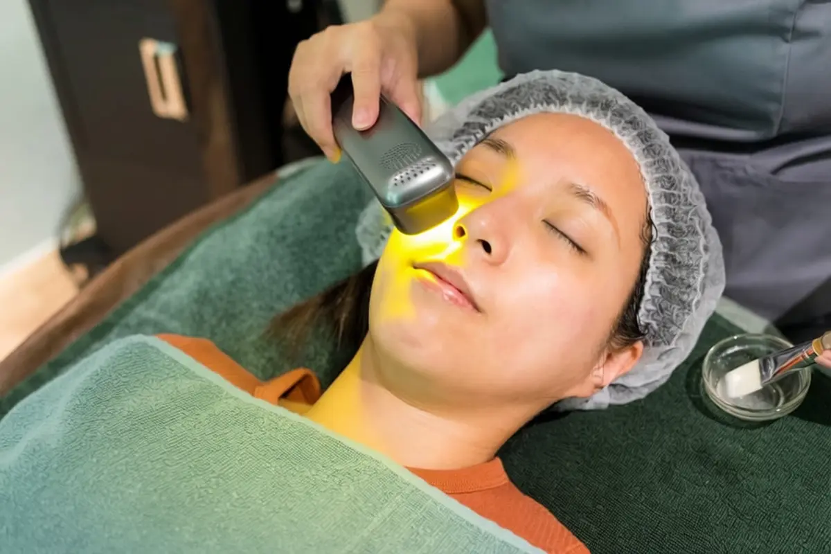 Yellow Laser Therapy Session - Before and After Treatment Care Tips Woman lying down with hair net and closed eyes receiving yellow laser therapy on her face showing visible yellow light on cheek and nose area