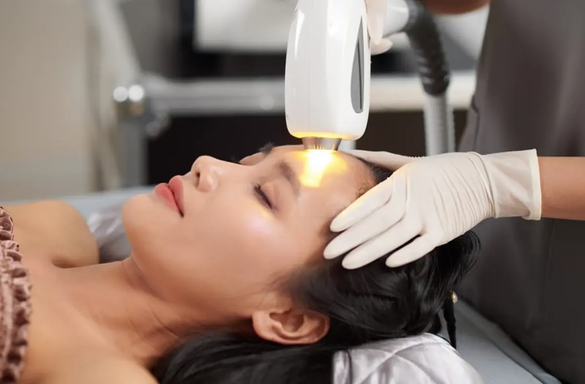 Woman receiving yellow laser treatment on face with eyes closed while practitioner applies handheld laser device emitting warm yellow light